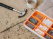 Top view of assorted repair instruments and screws in box near hammer and scotch tape on dirty tile in soft daylight