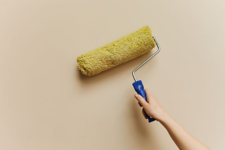 what to fix before selling a house — A hand using a yellow paint roller to paint a beige wall. Perfect for DIY and home improvement themes
